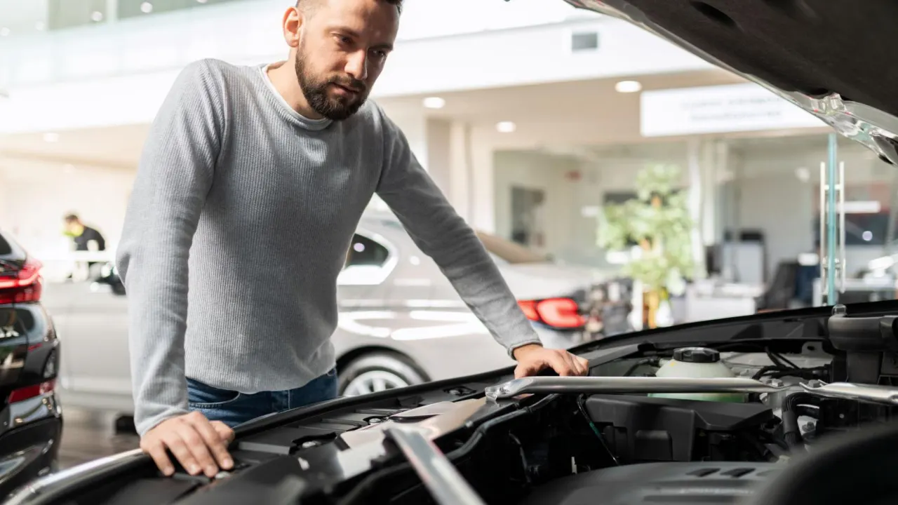 A man in a gray sweater inspects the engine bay of a modern vehicle indoors, possibly diagnosing car modules for engine or electronic faults.